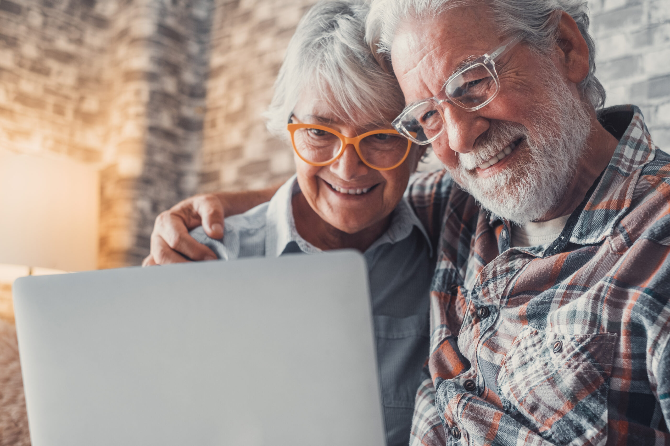 Cute couple of old people sitting on the sofa using laptop together shopping and surfing the net. Two mature people wearing eyeglasses in the living room enjoying technology. Portrait of seniors laughing in love.
